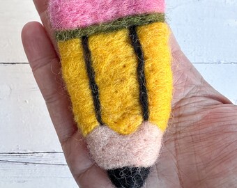Puede incluir: Un lápiz de fieltro con una goma de borrar rosa, cuerpo amarillo y líneas negras. El lápiz está sostenido en una mano.