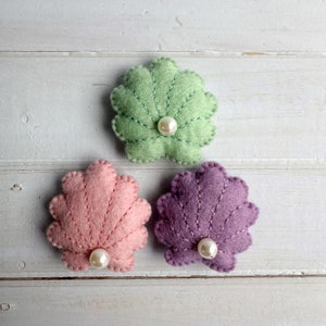 May include: Three felt seashell ornaments in pastel shades: light green, pink, and purple. Each shell features white stitching and a pearl bead at the centre. The ornaments are displayed on a white wooden surface.