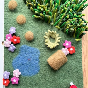 May include: A felt landscape scene with a green background, featuring a blue pond, brown spheres, and a cluster of green and yellow felt grass. Pink, red, and purple felt flowers are scattered around the scene, along with a brown felt pot with flowers.
