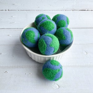 May include: A white bowl filled with seven felt balls, each depicting a blue and green globe. The bowl is sitting on a white wooden surface.