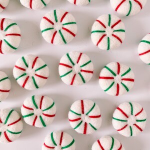 May include: A collection of felt peppermint ornaments in white with red and green stripes. The ornaments are arranged in a pattern on a white surface.