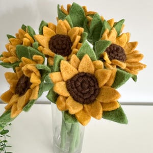 May include: A bouquet of felt sunflowers in a clear glass vase. The sunflowers have yellow petals and brown centres, with green felt leaves and stems. The arrangement is set against a white background.