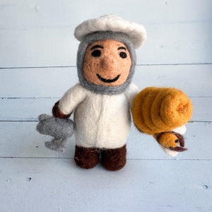 May include: A felted baker doll wearing a white chef's coat and hat, with a gray hood and beard. The doll holds a gray rolling pin in one hand and a golden-brown loaf of bread in the other. The doll has a smiling face.