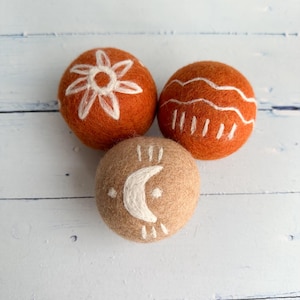 Peut inclure: Trois boules en feutre avec des motifs peints en blanc. Deux sont orange avec un motif de soleil et de vagues, et une est beige avec un motif de croissant de lune. Les boules sont disposées sur une surface en bois bleu clair.