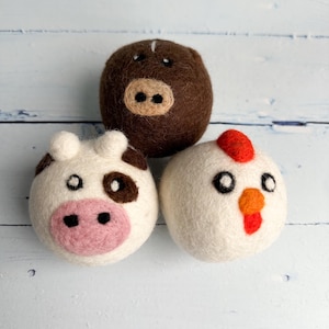May include: Three handmade felt animal ornaments: a brown pig, a white cow with brown patches, and a white chicken with a red comb and orange beak. The ornaments are round and have simple facial features.