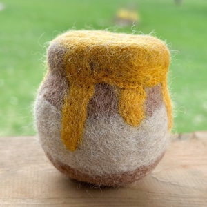 May include: A small, hand-felted honey pot with a brown and white base and a yellow felt top. The honey is dripping down the sides of the pot.