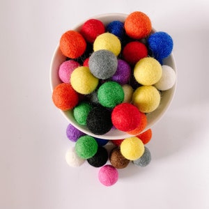 Könnte beinhalten: Eine weiße Schüssel gefüllt mit bunten Filzkugeln. Die Kugeln sind in verschiedenen Schattierungen von Rot, Orange, Gelb, Grün, Blau, Lila und Grau. Einige der Kugeln fallen aus der Schüssel.