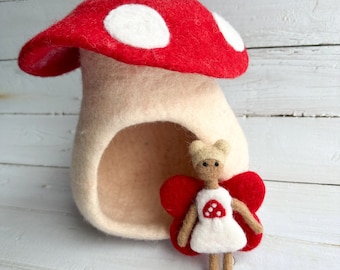 May include: A whimsical felt mushroom house with a red cap and white spots. A small doll with red butterfly wings and a mushroom design on its dress stands in front of the house. The house is beige and has an opening.