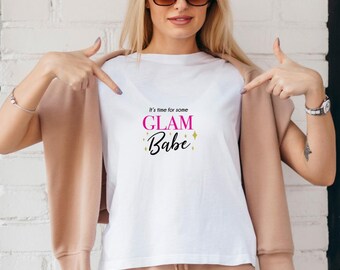 May include: White t-shirt with the text "It's time for some GLAM Babe" in pink and black. The shirt is being pointed at by a person wearing sunglasses and a tan jacket.
