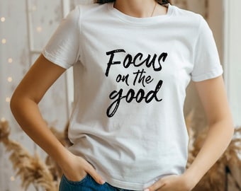 May include: White t-shirt with the phrase "Focus on the good" in black script. The shirt has short sleeves and a crew neck. The model is wearing blue jeans.