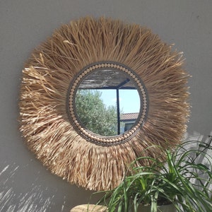 Peut inclure: Miroir mural rond avec un design en forme de soleil en raphia naturel. Le miroir a une bordure perlée et une garniture en corde tressée. Il est accroché à un mur gris clair. Un chapeau de paille et une plante en pot sont au premier plan.