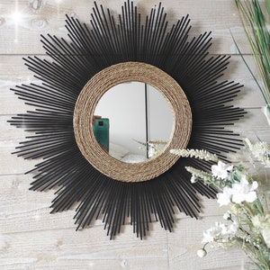 Op de afbeelding: Een ronde spiegel met een zonnestralenontwerp. De spiegel is omgeven door zwart geverfde houten stokjes en een frame van natuurlijke gevlochten touw. De spiegel hangt aan een witte houten muur.