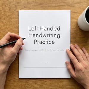 Könnte beinhalten: Ein weißes Übungsheft mit dem Titel Left-Handed Handwriting Practice, dem Untertitel 36 practice pages, built left-first - for teens and adults und einer Zeile für einen Namen. Ein schwarzer Stift liegt auf der Seite und eine weiße Tasse mit schwarzem Kaffee steht daneben.
