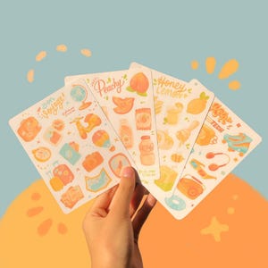 May include: A hand holds five sticker sheets with illustrations of travel, food, and tech themes. The sheets feature phrases like "Bon Voyage!" and "Honey Lemon." The stickers are in shades of orange, yellow, and blue.