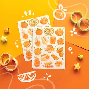 May include: Two sticker sheets featuring orange-themed illustrations, including oranges, juice, and macarons. The stickers have the text "ORANGE you glad". Decorative washi tape and star-shaped origami are also present, all in shades of orange and yellow.