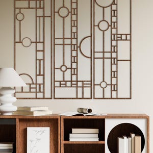 Könnte beinhalten: Dreiteilige Wandkunst mit geometrischem Design. Die Kunst zeigt eine Reihe von Rechtecken, Kreisen und Linien in brauner Ausführung. Das Kunstwerk ist an einer neutralfarbenen Wand über einem Bücherregal aus Holz mit Büchern montiert.