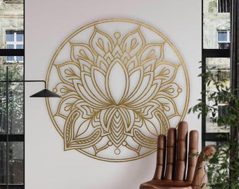 May include: A gold-colored lotus flower wall art piece is mounted on a white wall. The lotus design is circular and detailed. A wooden hand-shaped chair is positioned to the right of the wall art. The room has a modern aesthetic.