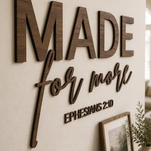 May include: A wall-mounted sign featuring dark brown wooden lettering that reads "MADE for more" in a combination of block and cursive fonts, with the text "EPHESIANS 2:10" positioned directly underneath in a smaller, simple sans-serif font.