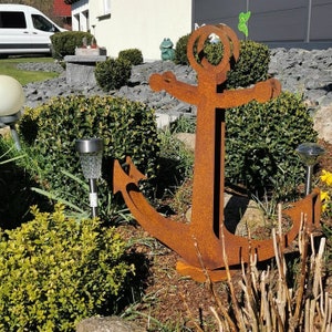 Könnte beinhalten: Eine große rostige Metallanker-Skulptur in einer Gartenlandschaft. Der Anker ist von grünen Büschen und braunem Mulch umgeben. Es gibt kleine Solarleuchten im Garten.