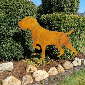 Könnte beinhalten: Eine rostige Metallsilhouette eines Hundes, der auf einem Stock steht, in einer Gartenumgebung. Der Hund blickt nach rechts im Bild. Der Hund ist von grünen Büschen und braunem Mulch umgeben. Im Vordergrund befinden sich mehrere hellbraune Steine.
