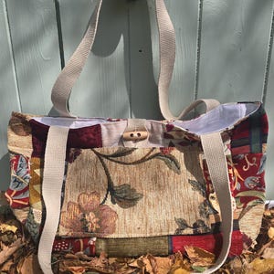 May include: A patchwork tote bag featuring a variety of floral and geometric fabric patterns in shades of red, gold, green, and cream. The bag includes two beige woven straps, a front pocket, and a wooden toggle closure with a beige fabric loop.