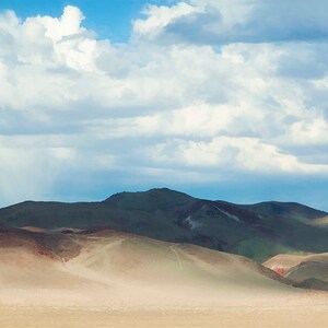 Może przedstawiać: Panoramiczny widok na pustynny krajobraz z falistymi wzgórzami i zachmurzonym niebem. Wzgórza są połączeniem brązu, zieleni i szarości, a niebo jest jasnoniebieskie z białymi chmurami.