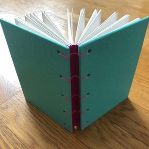 Op de afbeelding: Een teal linnen gebonden dagboek met een roze lint dat langs de ruggengraat is genaaid. Het dagboek is gesloten en ligt plat op een houten oppervlak.