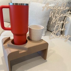 May include: A red insulated tumbler with a handle and a small white patterned cup sit in a light brown wooden drink holder. The holder has two circular cutouts. Text on the image reads "Available in: Walnut, Maple, Oak, Cherry."