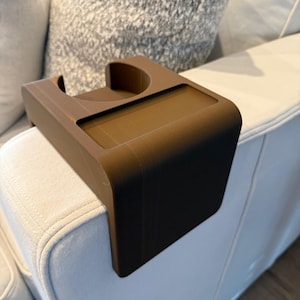 Puede incluir: Una bandeja marrón para el brazo del sofá con un portavasos circular y una bandeja rectangular. La bandeja está diseñada para colocarse en el brazo de un sofá. El producto está hecho de un material sólido y liso.