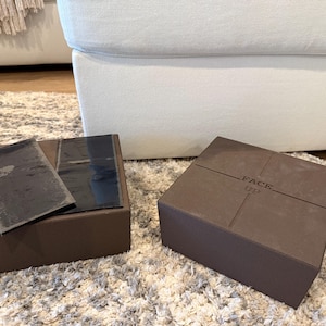 May include: Two brown square boxes, one open with black inserts, the other closed with "FACE UP" printed on the top. The boxes are on a light-colored rug, with a white sofa in the background.