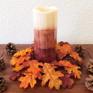 May include: A cylindrical pillar candle with a gradient color scheme transitioning from cream at the top to a deep burgundy at the base. The candle is surrounded by a ring of artificial autumn leaves in orange, yellow, and red, accented by six pinecones.