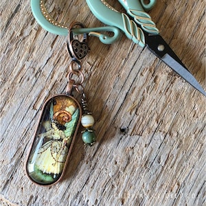 May include: A copper-toned pendant featuring an oval-shaped illustration of a winged figure in a light-colored dress, attached to a heart-shaped clasp with a small dangling charm consisting of a striped white bead and a green bead.