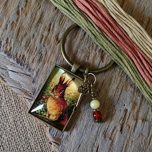 May include: A bronze-toned keychain featuring a rectangular pendant with a printed image of a brown rabbit surrounded by flowers. Attached to the ring is a small decorative skeleton key charm and two beads in pale yellow and deep red colors.