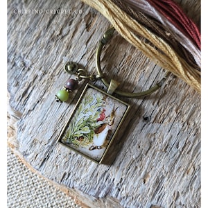 May include: A rectangular pendant featuring a printed illustration of a rabbit in a garden, set in a bronze-toned metal frame. The pendant hangs from a circular bronze ring adorned with a small heart charm and two beads in green and brown.