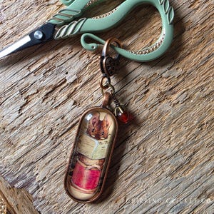 May include: A copper-toned oval pendant featuring an illustration of a mouse reading a newspaper while sitting on a red spool. The pendant hangs from a heart-shaped clasp attached to a pair of light green scissors with decorative metal detailing.