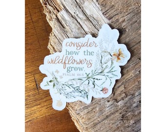May include: A die-cut sticker featuring the text consider how the wildflowers grow. PSALM 46:5 in brown and green script and serif fonts. The design includes watercolor-style illustrations of green stems, leaves, and small orange and white flowers.