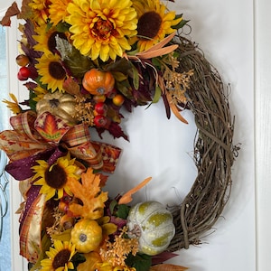 May include: A fall-themed wreath with a brown vine base, decorated with sunflowers, leaves, pumpkins, and berries. The wreath is adorned with a burgundy and gold plaid ribbon.