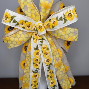 May include: A decorative bow featuring a sunflower and honeycomb pattern. The bow has multiple layers of yellow and beige ribbons. The center ribbon is white with a sunflower design. The bow is displayed on a white stand.