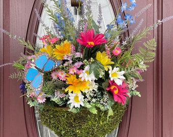 May include: A hanging floral basket overflowing with colorful artificial flowers, including pink, yellow, and white daisies, blue delphiniums, and lavender sprigs. A blue butterfly decorates the arrangement. The basket is covered in green moss and hangs on a brown door.