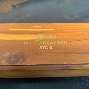May include: A rectangular wooden box with a natural wood grain finish. The box has gold lettering that reads "Smith's SOFT ARKANSAS STC-6". The box is closed and appears to be made of cedar wood.