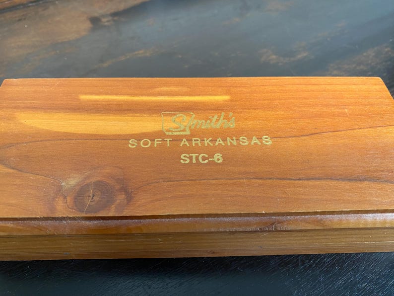 May include: A rectangular wooden box with a natural wood grain finish. The box has gold lettering that reads "Smith's SOFT ARKANSAS STC-6". The box is closed and appears to be made of cedar wood.