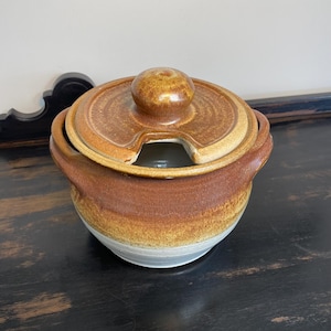 May include: A ceramic lidded serving bowl with a handle on each side. The bowl is a mix of brown, tan, and white, with a round knob on the lid. The lid has a notch for a spoon. The bowl is likely for serving soup or sauces.
