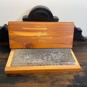 May include: A wooden box with a hinged lid, containing a gray sharpening stone. The lid is engraved with "Smith's SOFT ARKANSAS STC-6" in gold lettering. The box and stone are displayed on a dark wooden surface.
