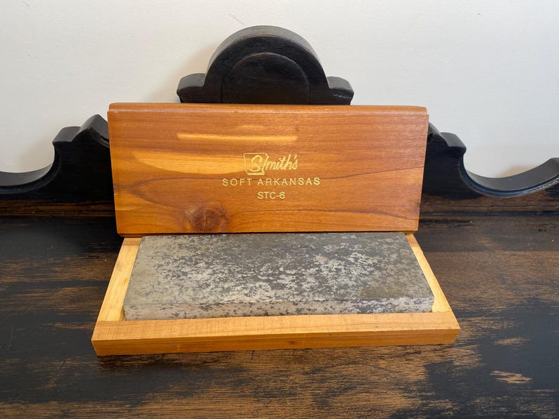 May include: A wooden box with a hinged lid, containing a gray sharpening stone. The lid is engraved with "Smith's SOFT ARKANSAS STC-6" in gold lettering. The box and stone are displayed on a dark wooden surface.
