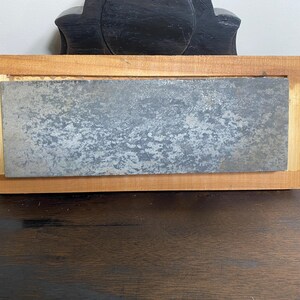 May include: A rectangular sharpening stone with a textured, gray surface is set within a light brown wooden frame. The wood frame has a lighter colored wood border. The item is designed for sharpening tools.