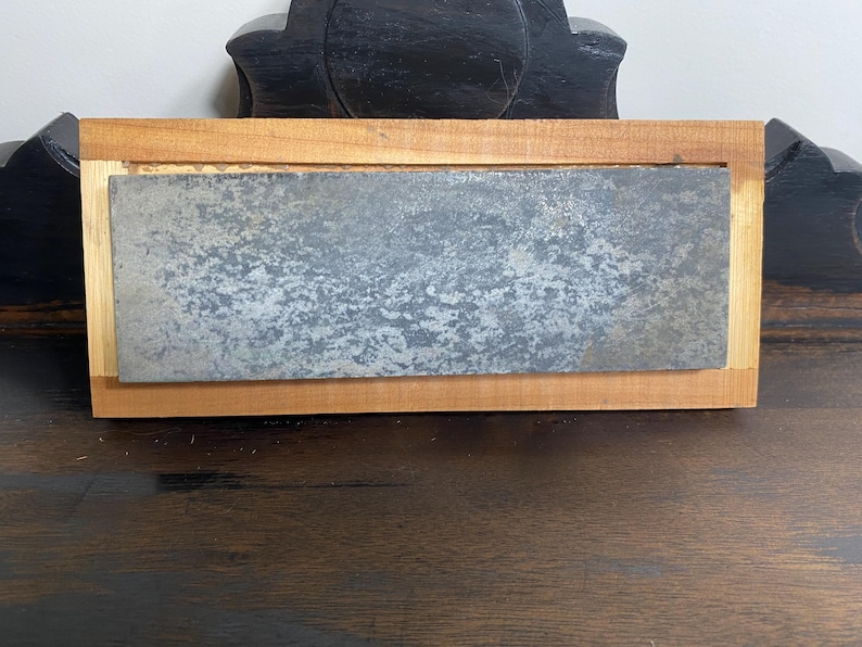 May include: A rectangular sharpening stone with a textured, gray surface is set within a light brown wooden frame. The wood frame has a lighter colored wood border. The item is designed for sharpening tools.