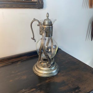 May include: An antique silver-toned glass carafe with a decorative handle and ornate base. The carafe has a clear glass body and a silver-toned lid with a small finial. The design includes decorative metalwork around the neck and base.