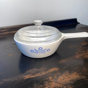 May include: A white ceramic saucepan with a single handle and a clear glass lid. The exterior of the pan features a blue floral pattern consisting of small flowers and leaves centered on the side. The lid has a small clear glass knob handle.