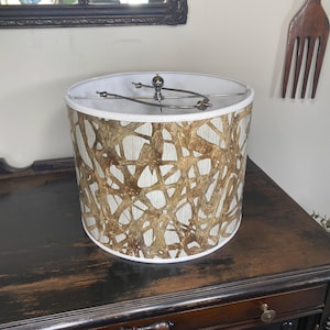 May include: Cylindrical lampshade with a white interior and a patterned exterior. The exterior features a network of brown, organic shapes against a light beige background. The lampshade has a metal frame and sits on a dark wooden surface.