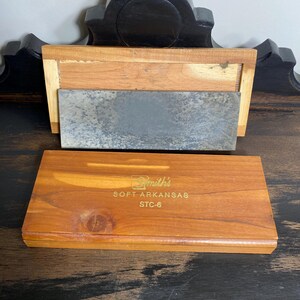 May include: A wooden sharpening stone set with a rectangular, gray stone and a wooden base. The set includes a separate wooden box with the text "Smith's SOFT ARKANSAS STC-6" in gold lettering. The wood has a warm, reddish-brown tone.